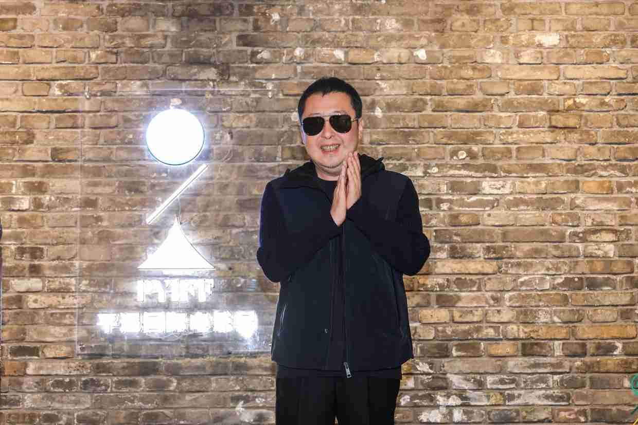 Man in sunglasses and a dark vest, smiling with hands clasped, stands before a brick wall with a glowing neon sign.
